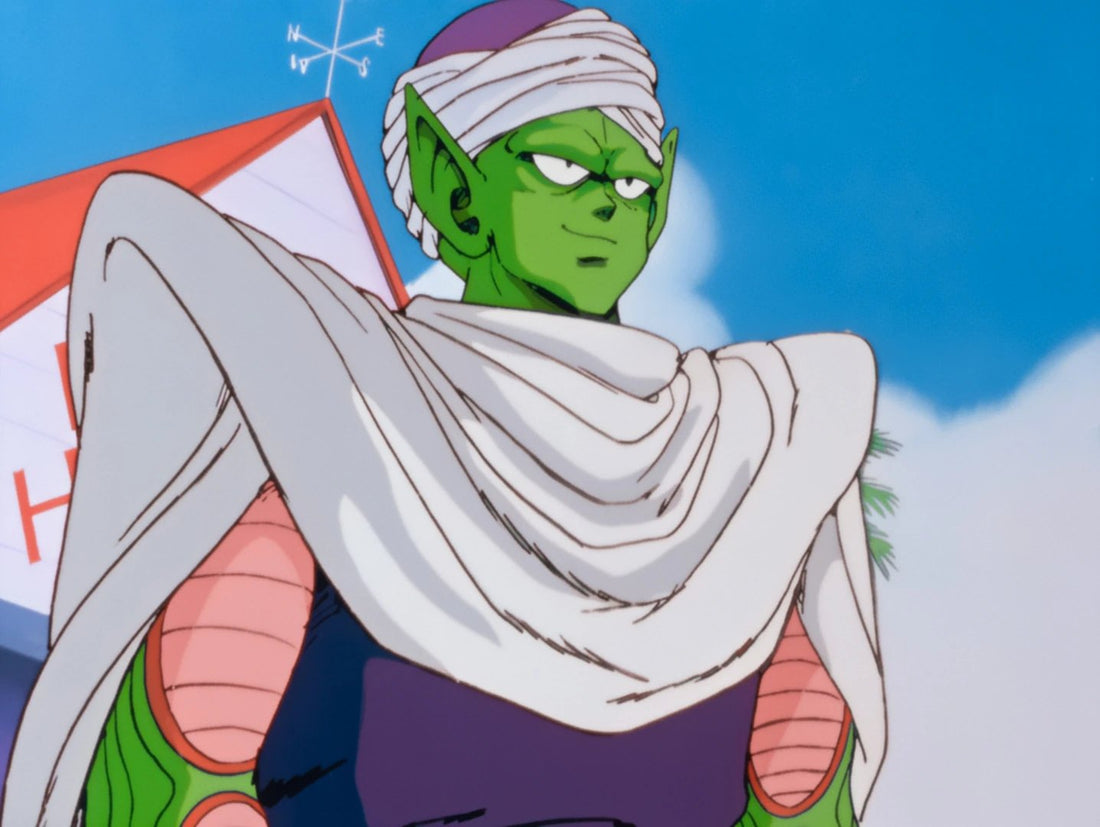 Piccolo, peau verte, turban blanc et grande cape blanche, sourit en coin sous un ciel bleu, devant un toit rouge surmonté d'une girouette.
